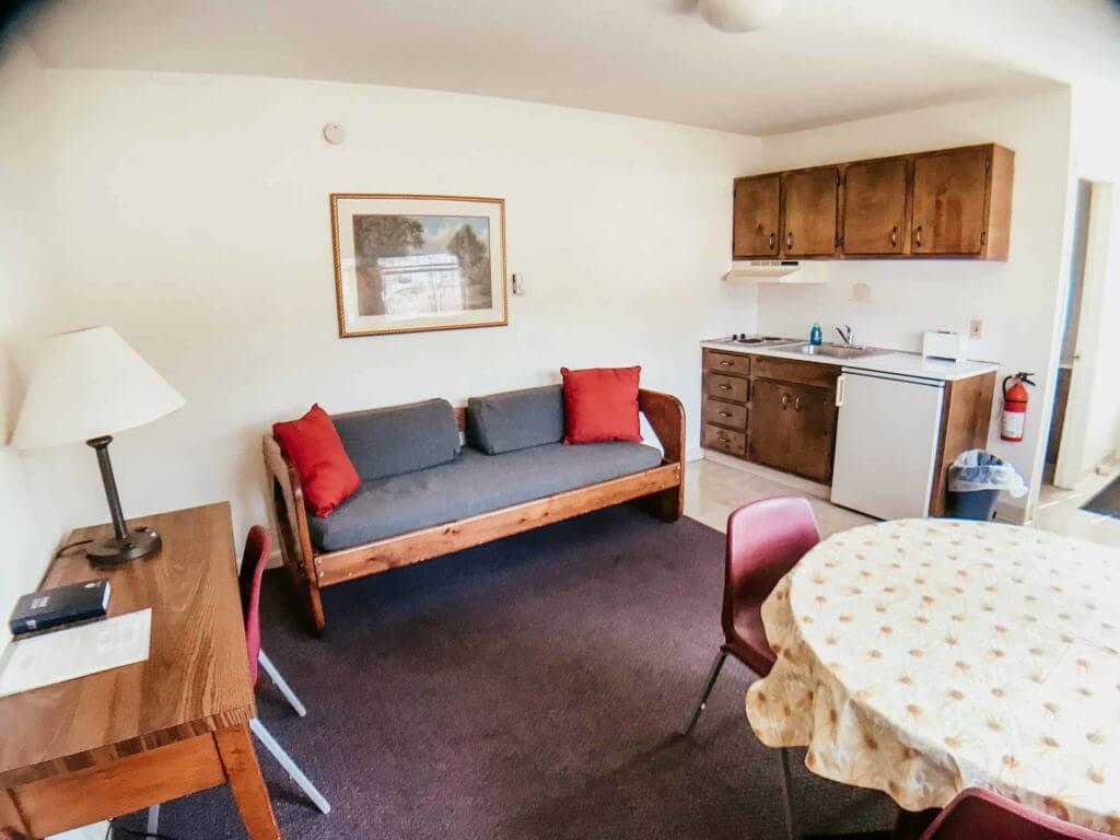 A small, simple living space designed for a christian retreat, featuring a wooden sofa with red cushions, a table with a yellow-patterned cloth, a wooden desk with lamp, and a kitchenette with brown cabinets, fridge, microwave, and fire extinguisher.