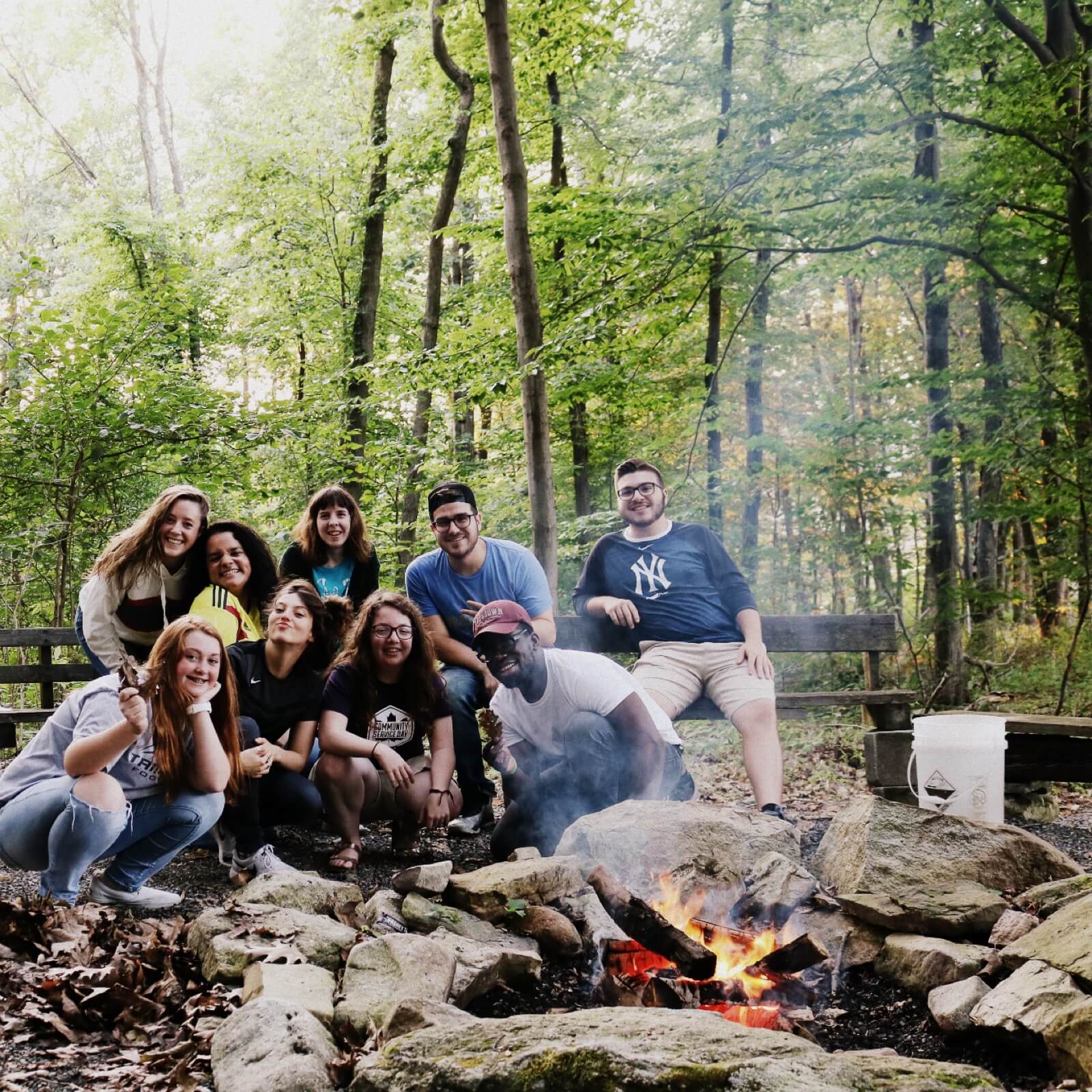 A group of nine young adults poses and smiles around a small campfire surrounded by rocks in a wooded area during a christian retreat, with green trees and a bench in the background.