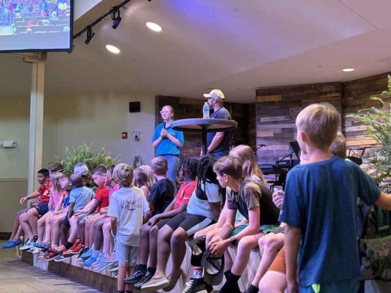 A man giving a speech to a live audience while children are seated and listening attentively.