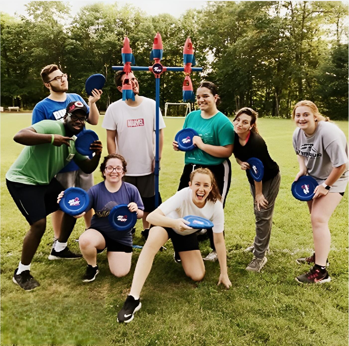 A group of eight people, smiling and holding blue frisbees, pose together in front of a frisbee game set on a grassy field with trees in the background during a christian retreat.