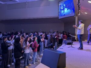 A man speaks on stage to a crowd in a large indoor auditorium during a christian retreat. Many in the audience stand near the stage, listening intently, while a screen above displays the stage scene.